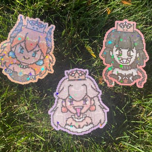 Peut inclure: Trois autocollants pixélisés d'une princesse avec des couronnes, dans des tons orange, noir et violet, avec des accents de paillettes. Les autocollants sont sur un lit d'herbe verte.