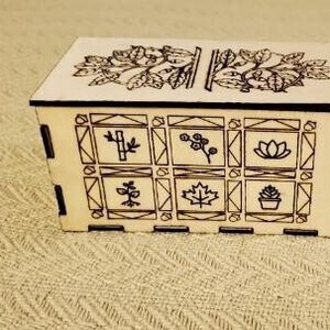 May include: A wooden box with a decorative design featuring a tree and various plant motifs. The box has a hinged lid and is made of light-colored wood.