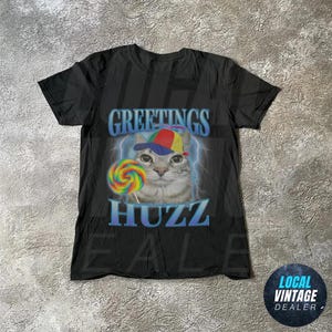 Op de afbeelding: Zwart t-shirt met een kat die een kleurrijke pet draagt en een lolly vasthoudt. De woorden "GREETINGS" en "HUZZ" staan in blauw met bliksemaccenten. De kat draagt een kleurrijke pet en houdt een regenbooglolly vast.
