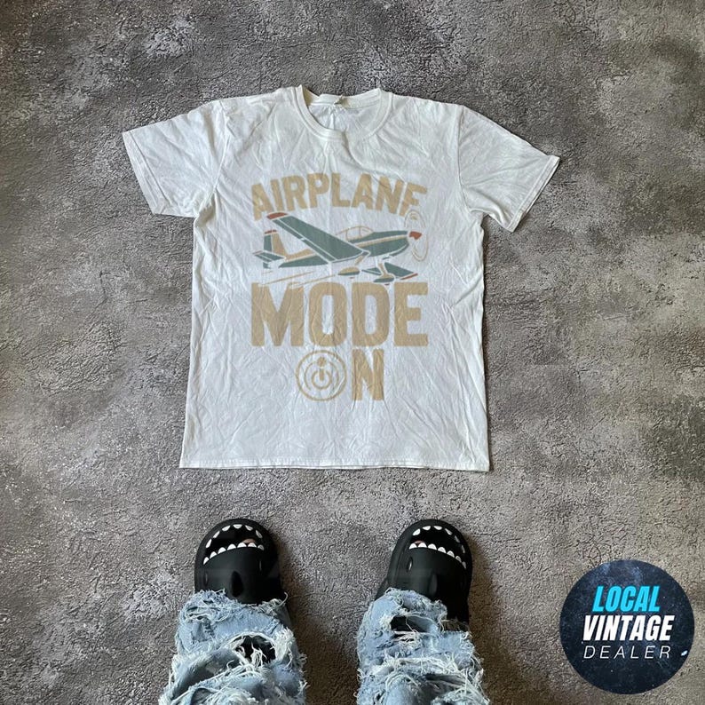 May include: Cream-coloured t-shirt featuring an airplane graphic and the text "AIRPLANE MODE ON" in beige. The airplane is teal, red, and white. The shirt is on a grey surface, with feet wearing shark-mouth shoes and ripped jeans.