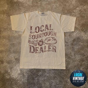 Könnte beinhalten: Cremefarbenes T-Shirt mit braunem Aufdruck "LOCAL SOURDOUGH DEALER" mit einer Brotlaib-Illustration. Das Shirt liegt flach auf einer strukturierten Oberfläche.