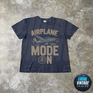 May include: Dark blue t-shirt featuring the text "AIRPLANE MODE ON" and a vintage airplane illustration. The design uses light blue and brown colours, with a propeller and the power button symbol.