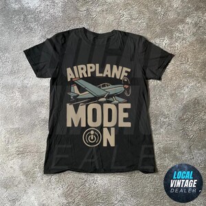 May include: Black t-shirt featuring a light blue airplane graphic and the words "AIRPLANE MODE ON" in a tan font. The design includes an airplane illustration and a power button symbol.
