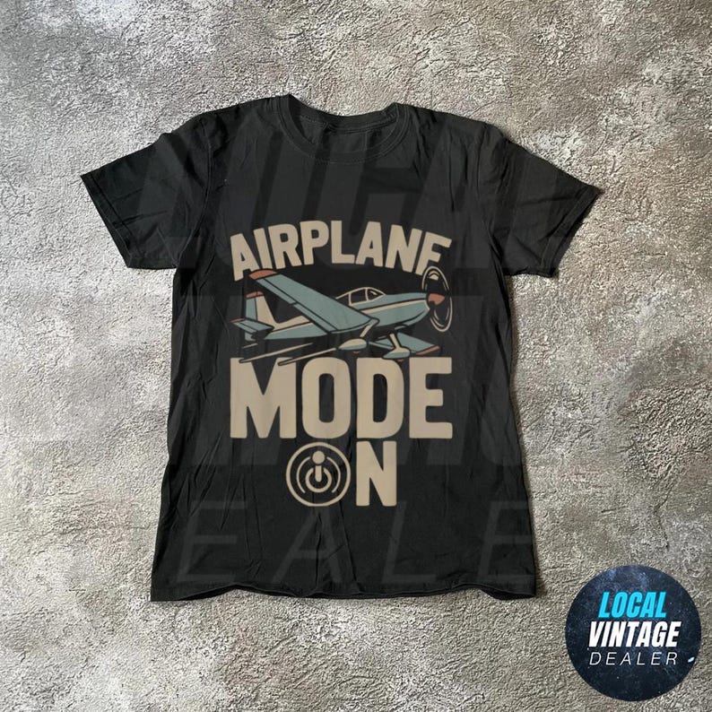 May include: Black t-shirt featuring a light blue airplane graphic and the words "AIRPLANE MODE ON" in a tan font. The design includes an airplane illustration and a power button symbol.