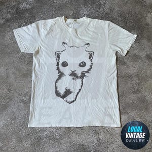 Op de afbeelding: Wit t-shirt met een zwart-witte illustratie van een katachtig wezen. De afbeelding staat op de voorkant van het shirt. Het shirt heeft korte mouwen en een ronde hals. Het shirt ligt op een grijze ondergrond.