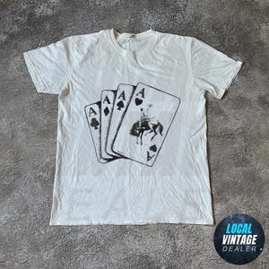 Könnte beinhalten: Weißes T-Shirt mit einer Grafik von vier Spielkarten, die jeweils ein Ass zeigen. Das Pik-Ass zeigt einen Cowboy auf einem Pferd. Das Shirt hat einen Rundhalsausschnitt.