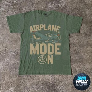 May include: A green t-shirt with the words "AIRPLANE MODE ON" in a vintage style. The shirt features a blue airplane graphic with red accents. The shirt is a crew neck and has short sleeves.