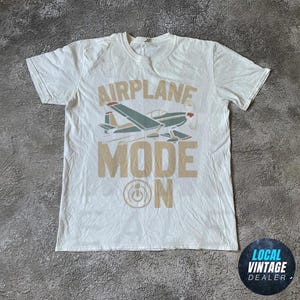 May include: White t-shirt featuring an airplane graphic and the text "AIRPLANE MODE ON" in tan. The airplane is teal, red, and tan. The shirt has a crew neck and short sleeves. The fabric appears slightly creased.