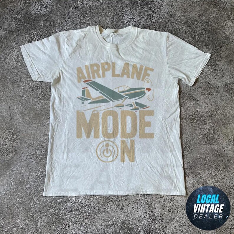 May include: White t-shirt featuring an airplane graphic and the text "AIRPLANE MODE ON" in tan. The airplane is teal, red, and tan. The shirt has a crew neck and short sleeves. The fabric appears slightly creased.