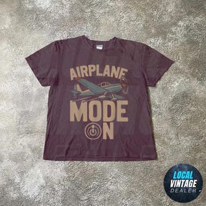 May include: A maroon t-shirt with a retro airplane graphic and the words "AIRPLANE MODE ON". The design features a blue and white aeroplane with a propeller. The shirt has a crew neck and short sleeves.
