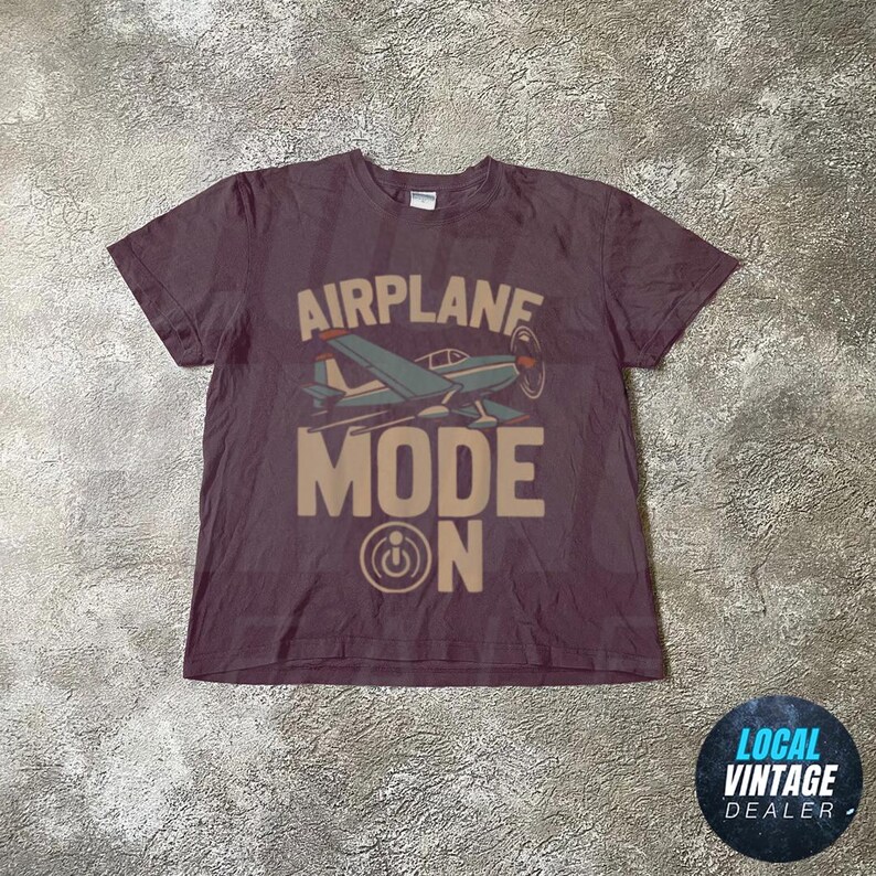 May include: A maroon t-shirt with a retro airplane graphic and the words "AIRPLANE MODE ON". The design features a blue and white aeroplane with a propeller. The shirt has a crew neck and short sleeves.