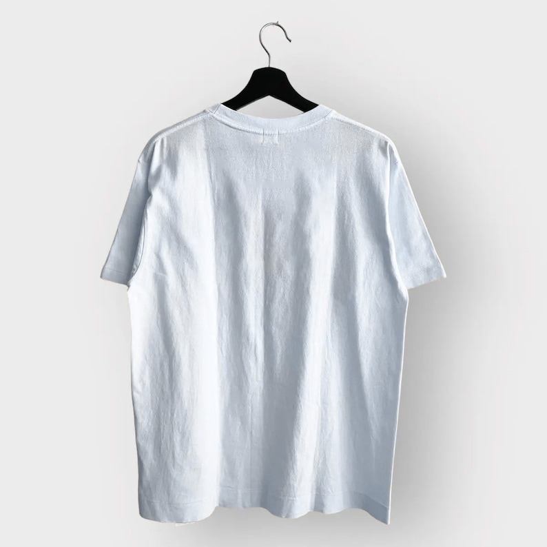 May include: A light blue t-shirt hangs on a black hanger against a white background. The short-sleeved shirt is made of a soft fabric and has a crew neck. The back of the shirt is visible, showing the simple design.