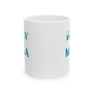 Op de afbeelding: Witte keramische mok met turquoise tekst die "KRAV MAGA" aan de ene kant en "KIM" aan de andere kant leest. De mok is cilindrisch met een gladde, glanzende afwerking, perfect voor warme dranken.