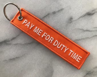 Puede incluir: Una etiqueta rectangular naranja para llavero con el texto bordado "PAY ME FOR DUTY TIME" en blanco. La etiqueta tiene un borde cosido y un llavero de metal plateado adjunto en la parte superior.