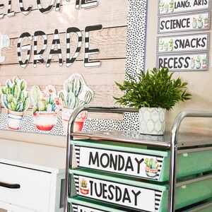 May include: A classroom display with the words "WELCOME TO 4 GRADE" in black and white lettering. Cactus cutouts and a schedule are also visible. A rolling cart with green bins labelled "MONDAY" and "TUESDAY" is in the foreground.