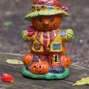May include: A ceramic scarecrow figurine with a pumpkin-shaped head and body. The scarecrow wears a yellow jacket, a red and yellow striped scarf, and a hat. Three carved pumpkins are at the base. The figurine is painted in bright colors.