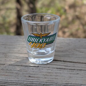 May include: Clear shot glass with the text "BRICKYARD 400" in green, gold, and white, with the date "August 1, 1998". The glass has a thick base and is sitting on a wooden surface.