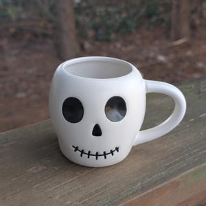 May include: A white ceramic mug with a black handle and the text "But First, Coffee" in a bold, black font. The mug is set against a blurred background, suggesting a cozy setting.