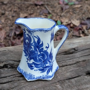 May include: A cream-colored ceramic pitcher featuring a hand-painted blue floral pattern. The vessel has a scalloped rim and base, both accented with a textured blue sponge-painted edge. A curved handle extends from the side of the body.