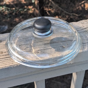 May include: A clear glass lid with a black knob. The lid is round and has a silver rim. The knob is a dark, matte black. The lid is sitting on a wooden surface.