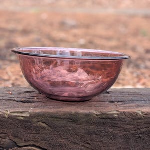 May include: A round, transparent, pink glass bowl with a black rim. The bowl is sitting on a weathered, dark brown wooden surface. The background is blurred, with hints of brown and green.