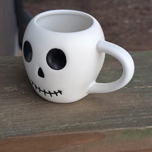 May include: White ceramic skull-shaped mug with black eye sockets, a nose, and a stitched mouth design. The mug has a rounded shape and a curved handle. The mug is sitting on a wooden surface.