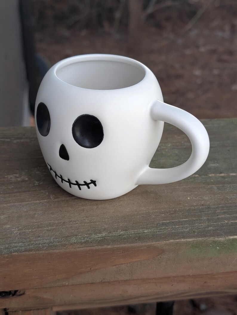 May include: White ceramic skull-shaped mug with black eye sockets, a nose, and a stitched mouth design. The mug has a rounded shape and a curved handle. The mug is sitting on a wooden surface.