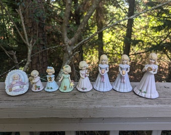 Op de afbeelding: Een set porseleinen beeldjes die een babymeisje voorstellen dat opgroeit. De beeldjes zijn gekleed in pastelkleurige jurken en houden nummers vast die elk jaar van leeftijd voorstellen. De beeldjes zijn in een rij op een houten oppervlak gerangschikt.