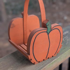 May include: Wooden pumpkin-shaped basket painted orange with a black outline and green stem. The basket has a handle and is designed with a slatted base. A seasonal decorative item for fall.