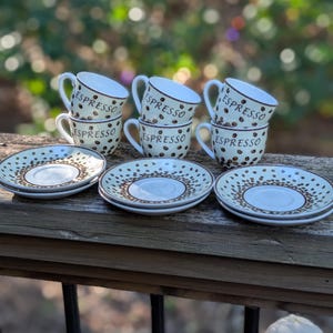 May include: A set of three white ceramic espresso cups and matching saucers, each decorated with a brown coffee bean pattern and the word ESPRESSO printed in black. The cups feature a curved handle and a white interior, while the saucers have a circular design with a matching coffee bean motif around the rim.
