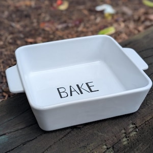 May include: A white square baking dish with the word "BAKE" in black lettering. The dish has two small handles and is sitting on a weathered wooden surface. The dish is empty.