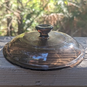May include: A brown glass lid with a knob on top. The lid has a ribbed pattern and is slightly translucent.