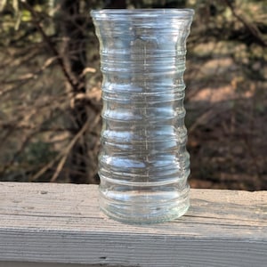 May include: A clear glass vase with a ribbed design. The vase has a cylindrical shape with a slightly flared top and base. The vase is sitting on a wooden surface, with a blurred background of trees.
