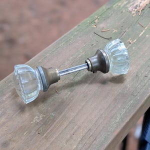 May include: A door knob set featuring two clear, faceted glass knobs connected by a central metal spindle. Each glass knob is attached to a circular, tarnished brass-toned metal base plate. The metal spindle is silver-toned and cylindrical.