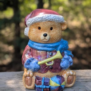 May include: A ceramic teddy bear figurine wearing a Santa hat, blue scarf, and gloves, holding a yellow trumpet. The bear is dressed in a red coat and has a stack of presents at its feet. The figurine is painted in various colors.