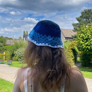 May include: A blue crochet bucket hat with a white trim. The hat features a gradient of blues, transitioning from dark navy to light blue. The hat is worn outdoors under a cloudy sky.