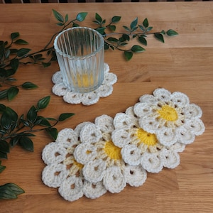 Könnte beinhalten: Vier gehäkelte weiße Untersetzer in Form von Gänseblümchen mit gelben Zentren und ein Glas auf einem weißen gehäkelten Untersetzer.