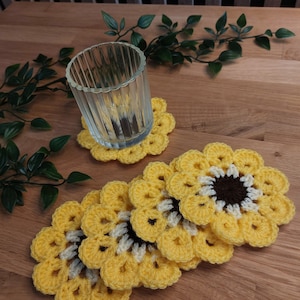 Könnte beinhalten: Satz von fünf gehäkelten Sonnenblumen-Untersetzern in Gelb, Weiß und Braun. Die Untersetzer sind auf einer Holzoberfläche gestapelt.