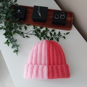 May include: A pink and coral knitted beanie hat. The hat is made of soft yarn and has a ribbed texture.