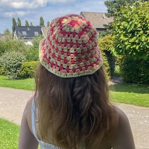 Könnte beinhalten: Ein gehäkelter Bucket-Hut in Rosa, Gelb und Weiß. Der Hut ist mit einem quadratischen Stichmuster gearbeitet.