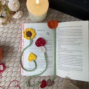Può includere: Segnalibro all'uncinetto con disegni di girasole, fungo e cuore in giallo, rosso e marrone. Il segnalibro è posto su un libro aperto con testo in tedesco e una candela accesa sullo sfondo.