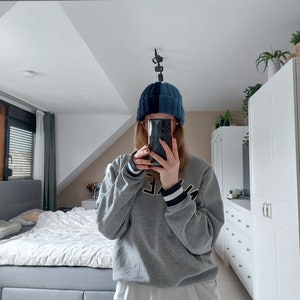 May include: A person wearing a gray sweatshirt with a white undershirt and black leggings. They are wearing a blue and black knit beanie and holding a black smartphone in front of their face.