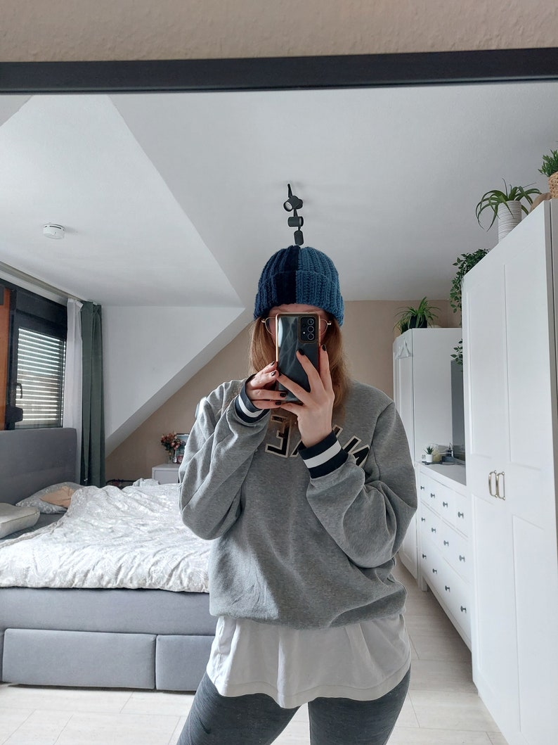 May include: A person wearing a gray sweatshirt with a white undershirt and black leggings. They are wearing a blue and black knit beanie and holding a black smartphone in front of their face.