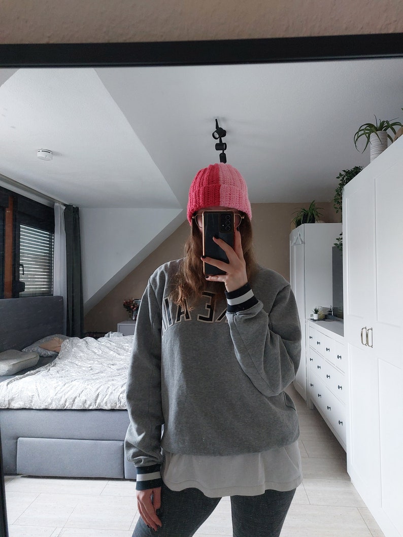 May include: A person wearing a gray sweatshirt with black and white stripes on the cuffs, black leggings, and a pink and red knit beanie. The beanie has a vertical stripe down the center.