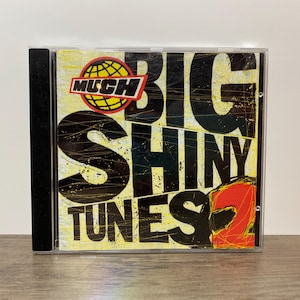 May include: A CD case with the album cover for "Big Shiny Tunes 2." The cover features the text "BIG SHINY TUNES 2" in large black letters over a yellow and black background. The MuchMusic logo is at the top.