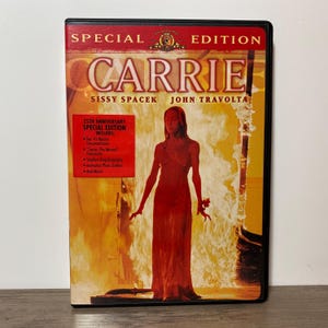 May include: A DVD case for the movie "Carrie" featuring Sissy Spacek and John Travolta. The cover art shows a woman in a red dress against a fiery background. The case is a special 25th-anniversary edition.