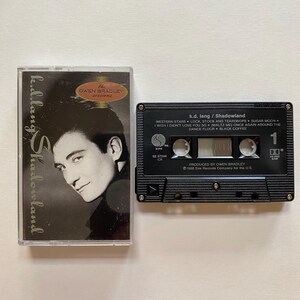 May include: A black cassette tape with the title "k.d. lang / Shadowland" and the text "WESTERN STARS * LOCK, STOCK AND TEARDROPS * SUGAR MOON * I WISH I DIDN'T LOVE YOU SO * WALTZ ME * ONCE AGAIN AROUND THE DANCE FLOOR * BLACK COFFEE" printed on the front. The cassette tape is next to a black and white photo of k.d. lang on a cassette tape case.