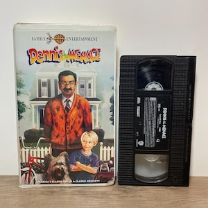May include: A VHS tape of the movie "Dennis the Menace" with the Warner Bros. Family Entertainment logo. The cover features a man, a boy, and a dog in front of a house. The tape is black.