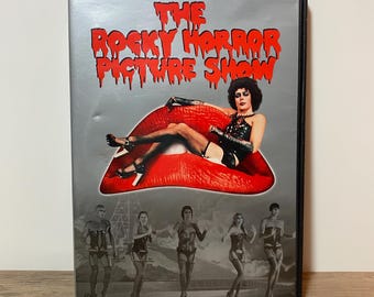 Puede incluir: Caja de DVD de "The Rocky Horror Picture Show" con fondo plateado. La portada presenta a una persona en lencería reclinada sobre un labio rojo, con el título de la película en letras rojas goteando. Debajo, una fila de bailarines en lencería.
