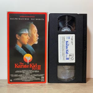 May include: A vintage VHS tape of "The Karate Kid Part II" movie. The cover features Ralph Macchio and Pat Morita. The black VHS tape is next to the cover, with the movie title and credits.